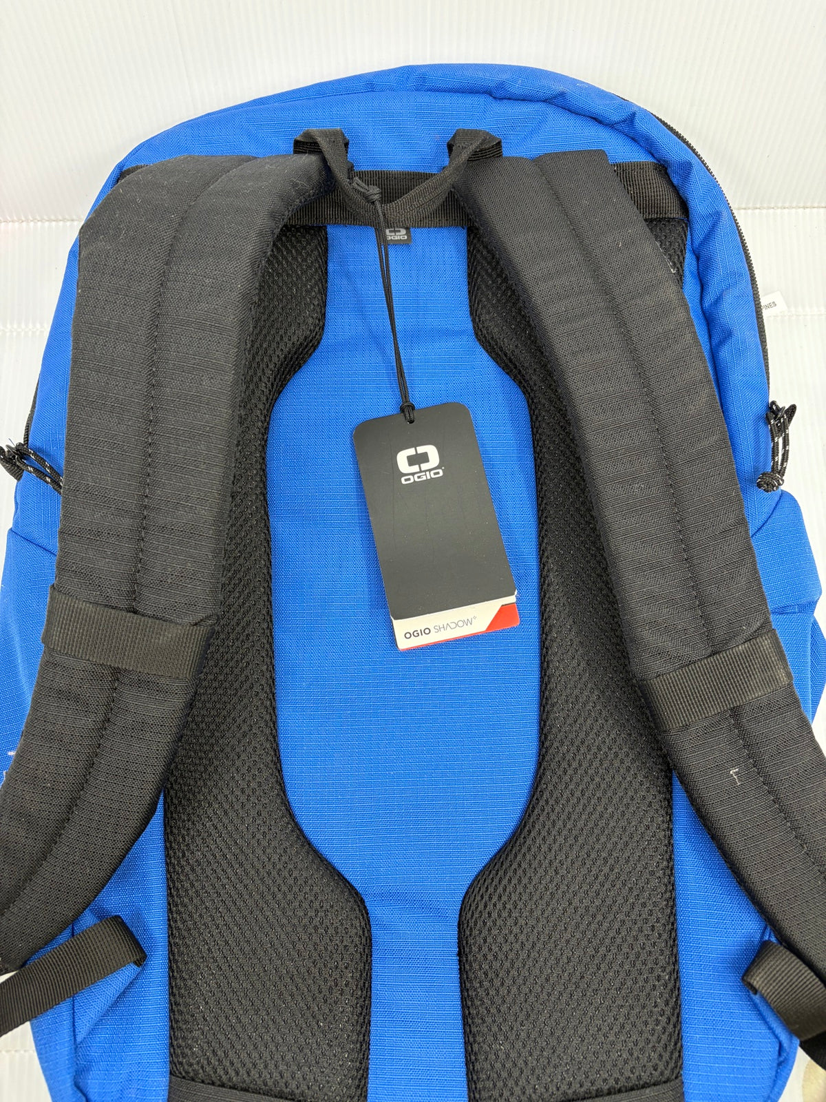 OGIO Shadow blue backpack back view with padded black shoulder straps, mesh back panel and OGIO Shadow hangtag