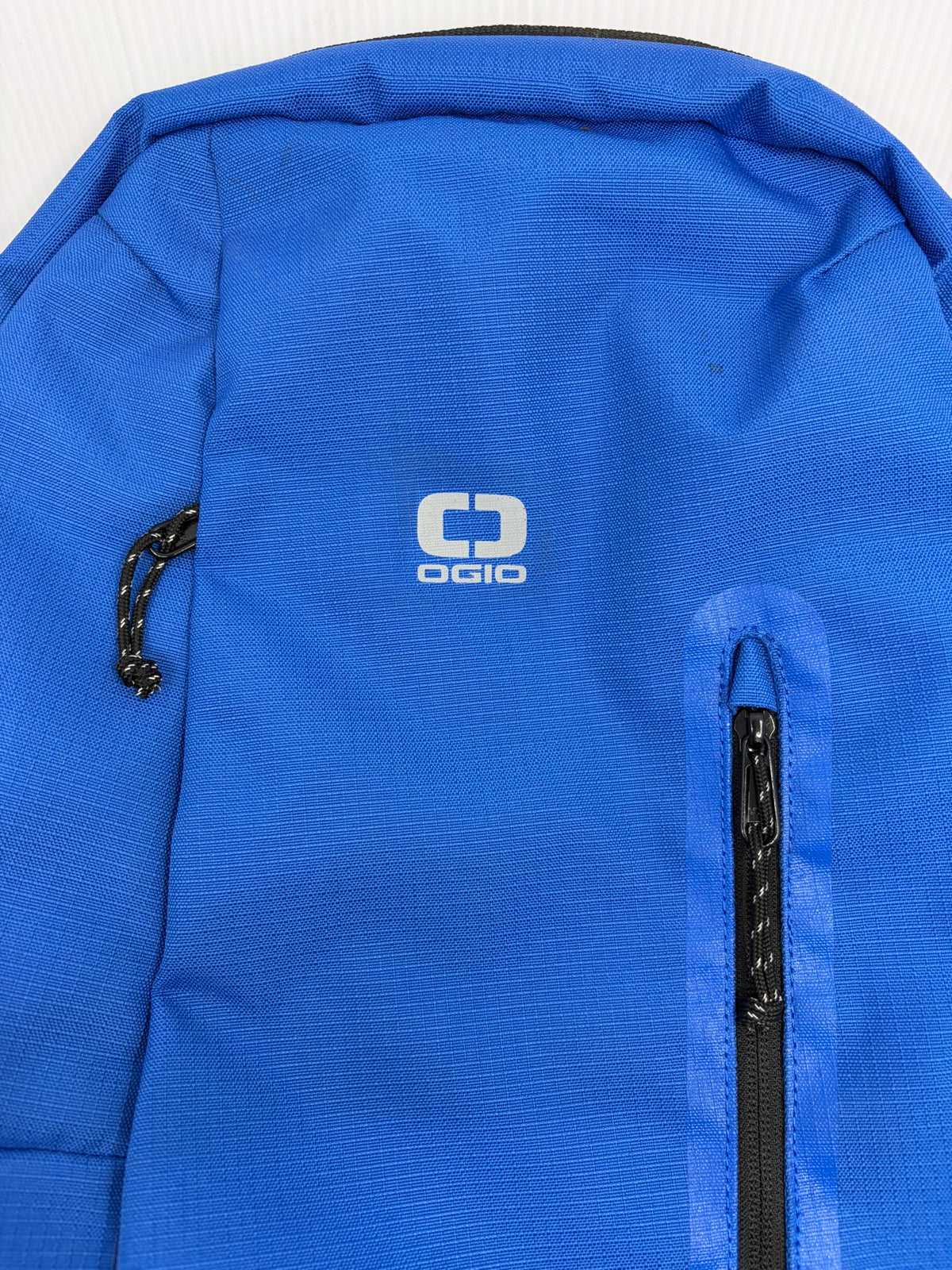 Close-up of blue ripstop fabric and white OGIO logo on the Shadow backpack front, showing tiny dark specks near the top