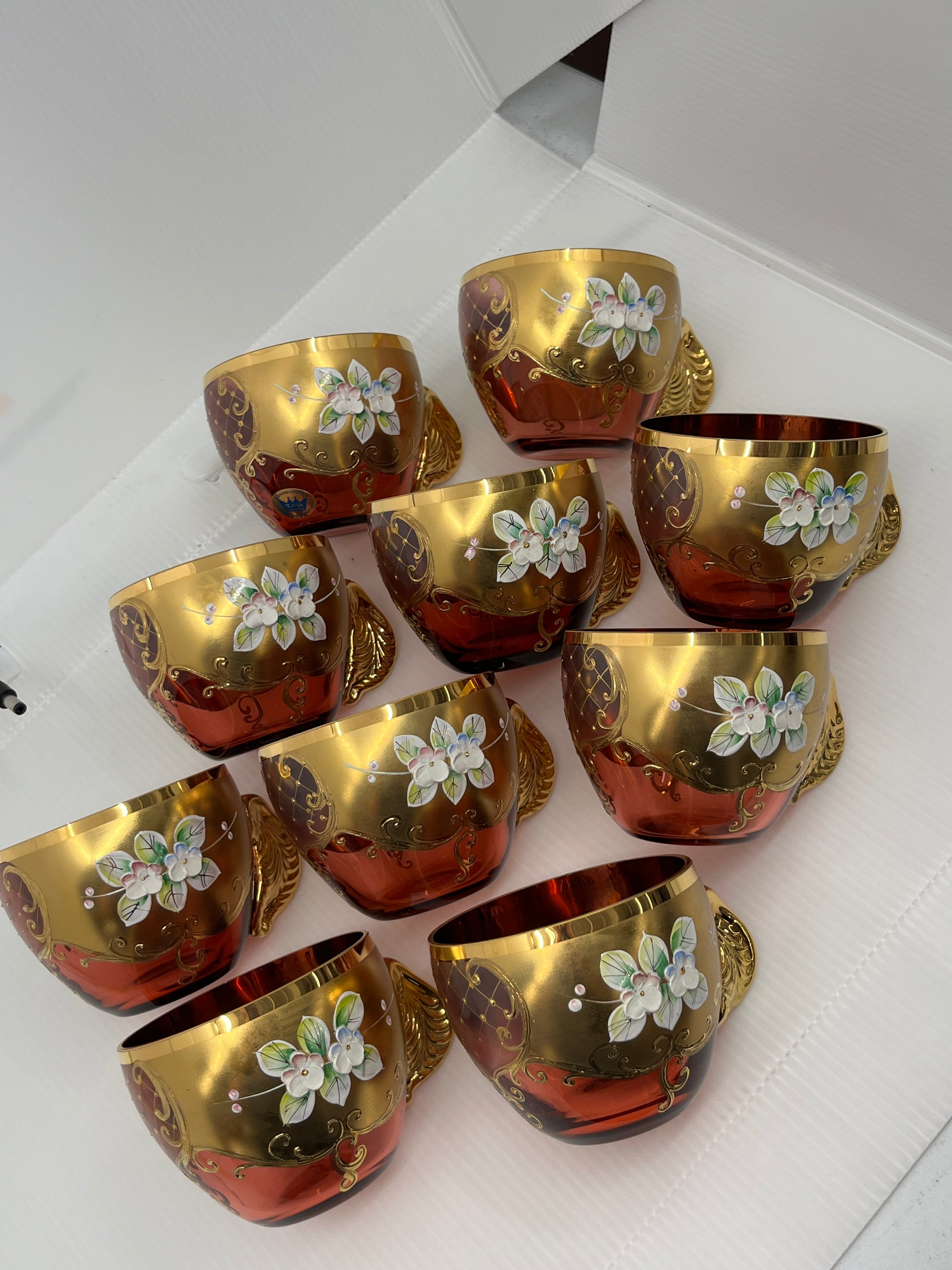 Ten gold and red punchbowl teacups with floral designs on a white surface.